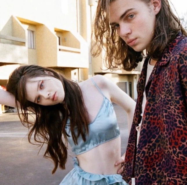 Young couple poses outdoors; woman in blue satin top and skirt, man in animal print jacket with apartment buildings in the background.