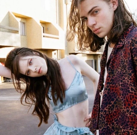 Young couple poses outdoors; woman in blue satin top and skirt, man in animal print jacket with apartment buildings in the background.