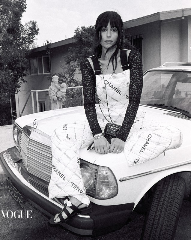 Black and white shot of a woman in a Chanel dress sitting on the hood of a classic car in front of a house and statue.