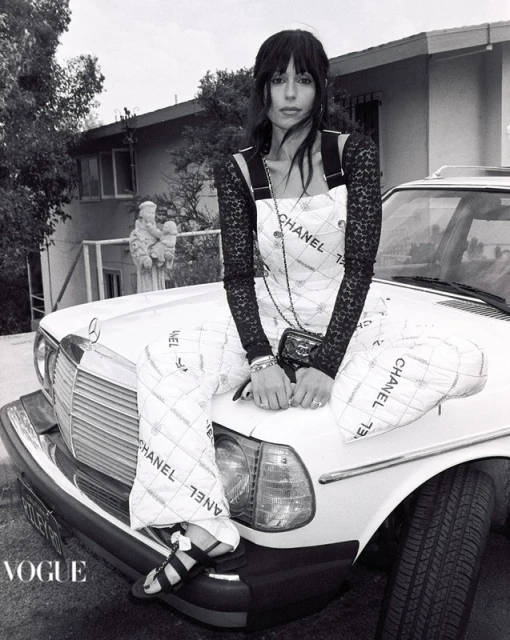 Black and white shot of a woman in a Chanel dress sitting on the hood of a classic car in front of a house and statue.