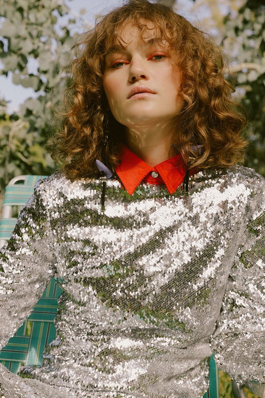 Woman with curly hair poses in sequin dress and red collar, seated outdoors, under soft sunlight and dappled tree shadows.