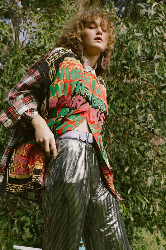 A woman in a patterned shirt and silver pants stands outdoors, surrounded by green foliage on a sunny day, showcasing fashion and style.