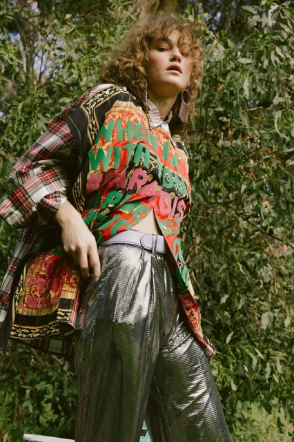 A woman in a patterned shirt and silver pants stands outdoors, surrounded by green foliage on a sunny day, showcasing fashion and style.