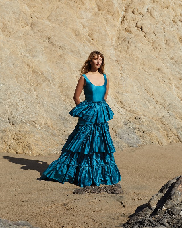 Woman in teal formal gown on a beach, posing in front of a rock formation. Fashion shot with natural light, warm tones, and coastal setting.