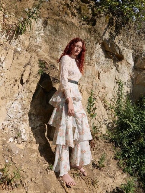 A red-haired woman in a tiered floral dress poses outdoors in front of a rocky cliff, gazing toward the camera in natural daylight.