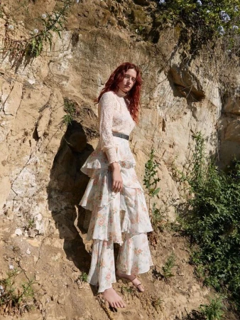 A red-haired woman in a tiered floral dress poses outdoors in front of a rocky cliff, gazing toward the camera in natural daylight.