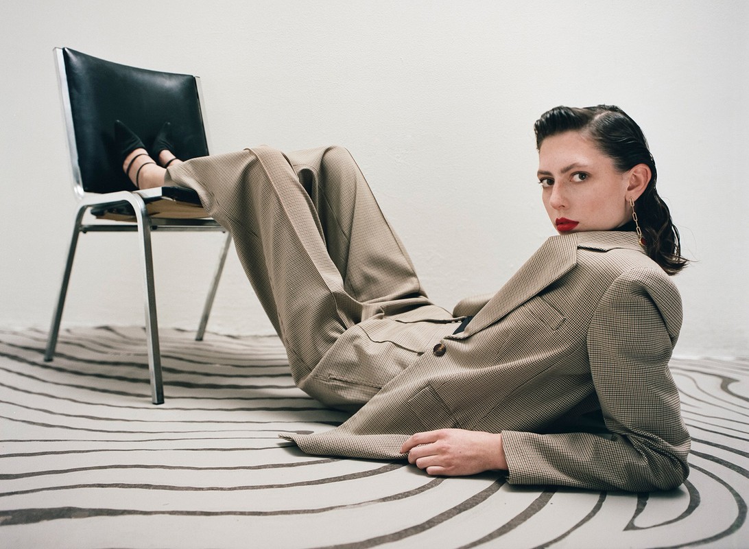 A woman in a tan suit reclines on a striped rug, her feet resting on a metal and black chair. She looks directly at the camera.