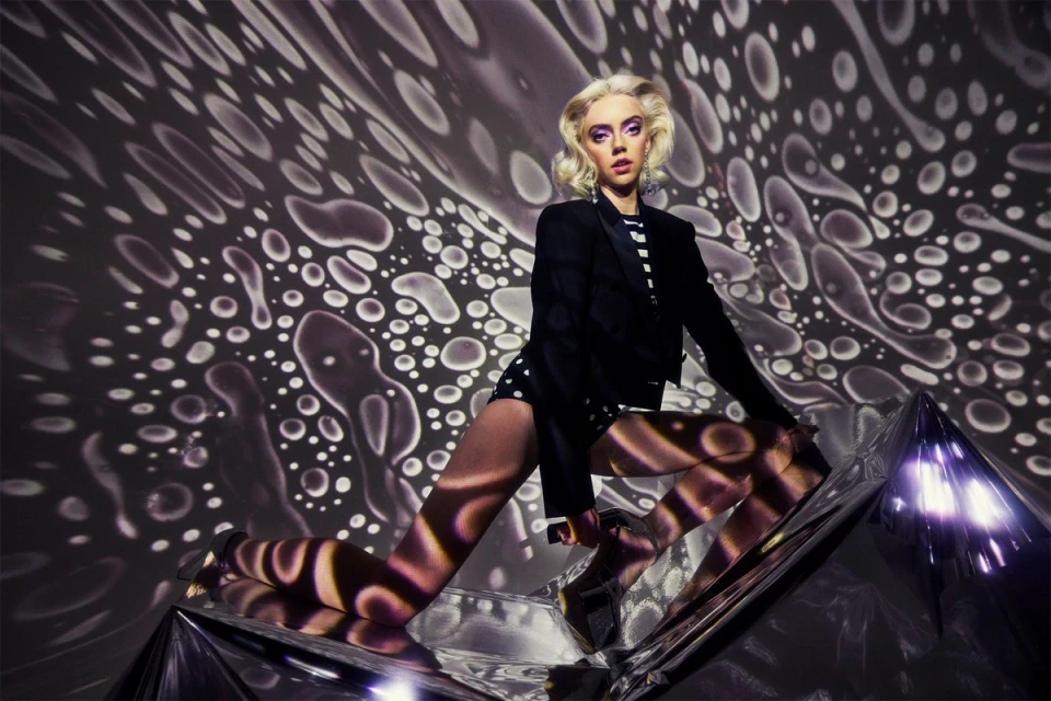 Portrait of blonde adult female in black blazer and polka dot shorts sitting on shiny metallic surface, background of white dots on black.