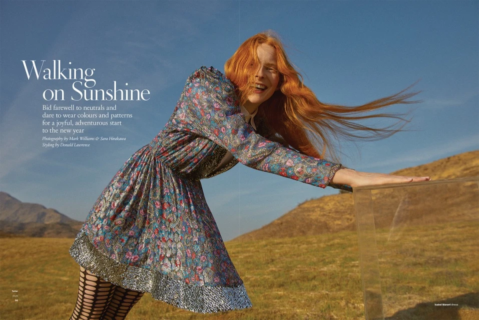 A red-haired fashion model smiles outdoors wearing a floral print mini dress, black fishnet tights, and leaning against a clear box.