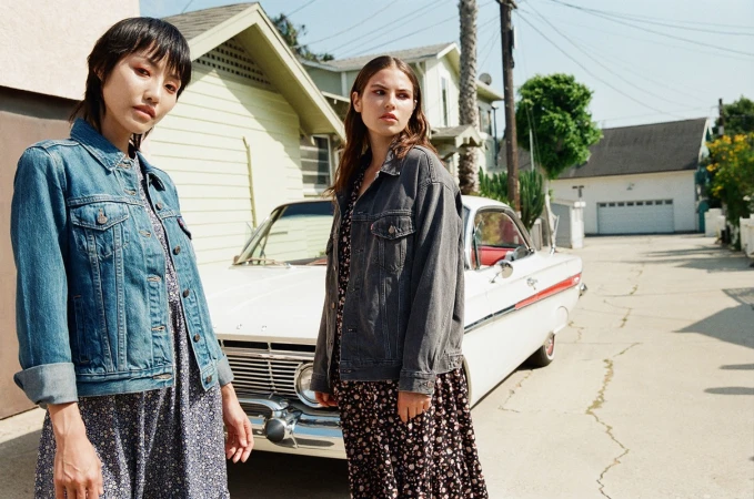 Two adult women in denim jackets and floral dresses pose in front of a vintage white car and suburban houses on a sunny day.