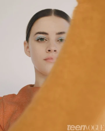 Close-up portrait of a woman with blue eyeshadow and dark hair parted centrally, partly obscured by an orange fabric.