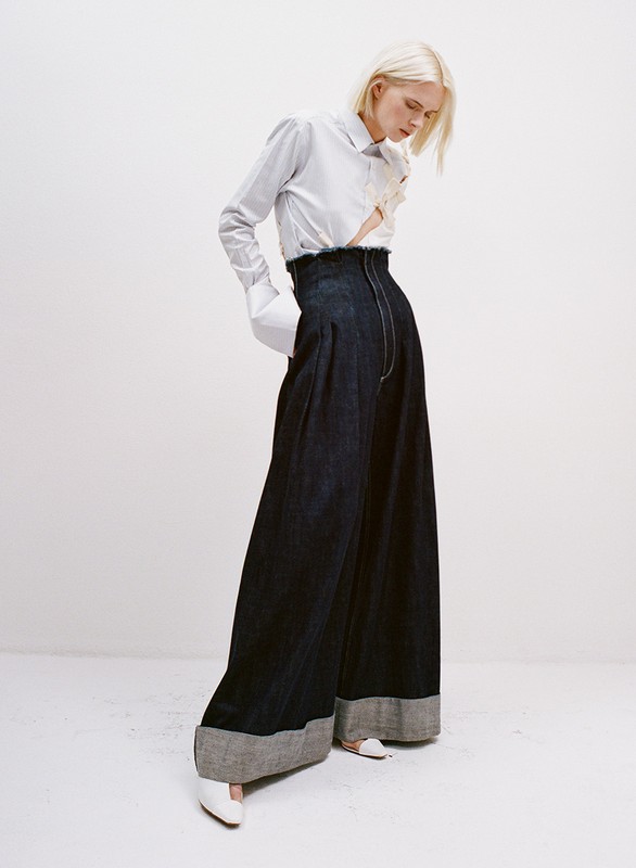 Fashion shot of a fair-skinned woman with blonde hair, wearing a striped blouse and dark, wide-legged denim pants in a white studio.
