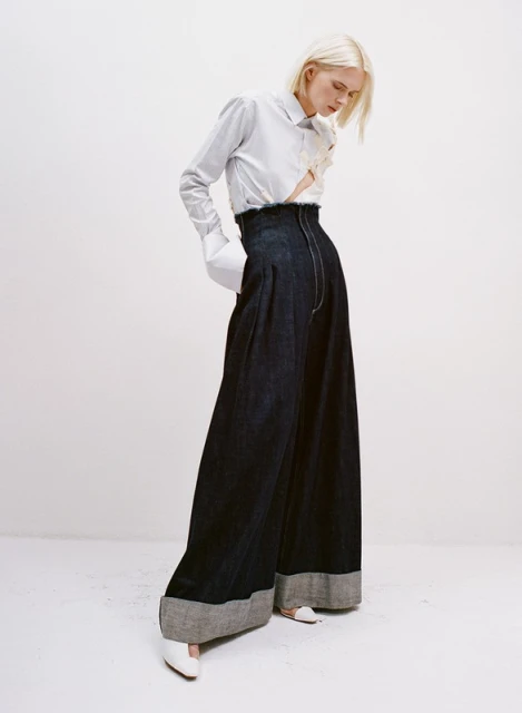 Fashion shot of a fair-skinned woman with blonde hair, wearing a striped blouse and dark, wide-legged denim pants in a white studio.