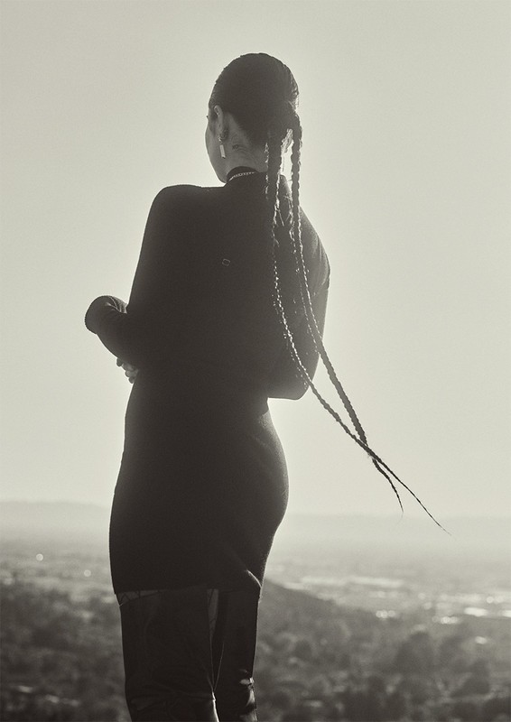 Woman seen from behind with long braided pigtails wearing a dark fitted dress against a bright sky and distant horizon