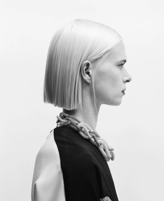 Monochrome profile shot of a woman with platinum blonde bob, wearing a chunky braided necklace, against a light background.