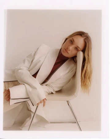 Portrait of a woman with long blonde hair wearing a white jacket over a burgundy top, seated and leaning against a chair.