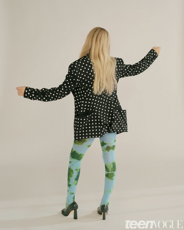 Back view of an adult female in a black polka dot blazer, green and blue patterned leggings, and green heels against a white background.