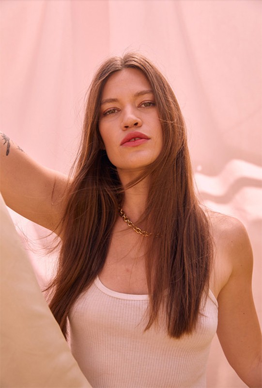 Portrait of a woman with long brown hair wearing a white tank top and gold chain necklace against a textured pink background.