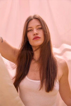 Portrait of a woman with long brown hair wearing a white tank top and gold chain necklace against a textured pink background.