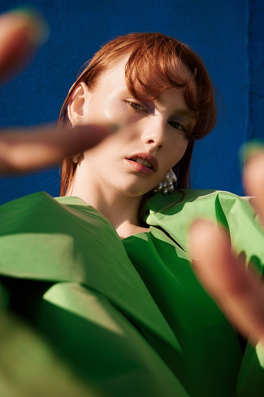 Close-up fashion shot of a red-haired model wearing a green sculptural garment against a blue backdrop, with hands reaching into frame.