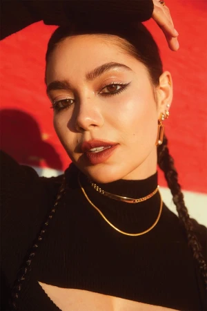 Close-up portrait of a woman with braided dark hair wearing gold chains and a black top against a red wall.