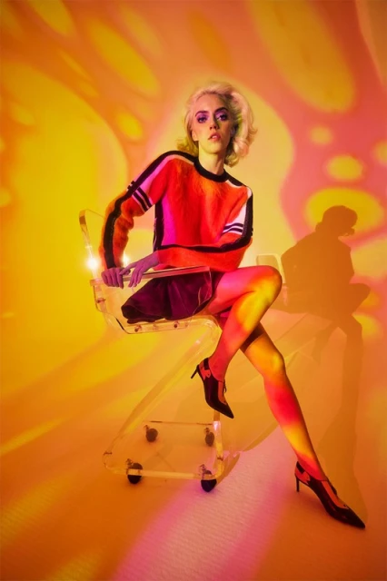 Stylish woman with blonde hair poses in front of an abstract orange background while seated in a transparent chair with her legs crossed.