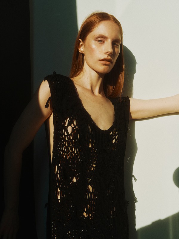 A red-haired woman poses in natural light wearing a black open-knit vest with a wall as background. Fashion editorial.