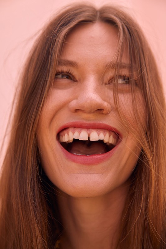 Close up portrait of a laughing woman with long brown hair and a visible tooth gap against a plain background