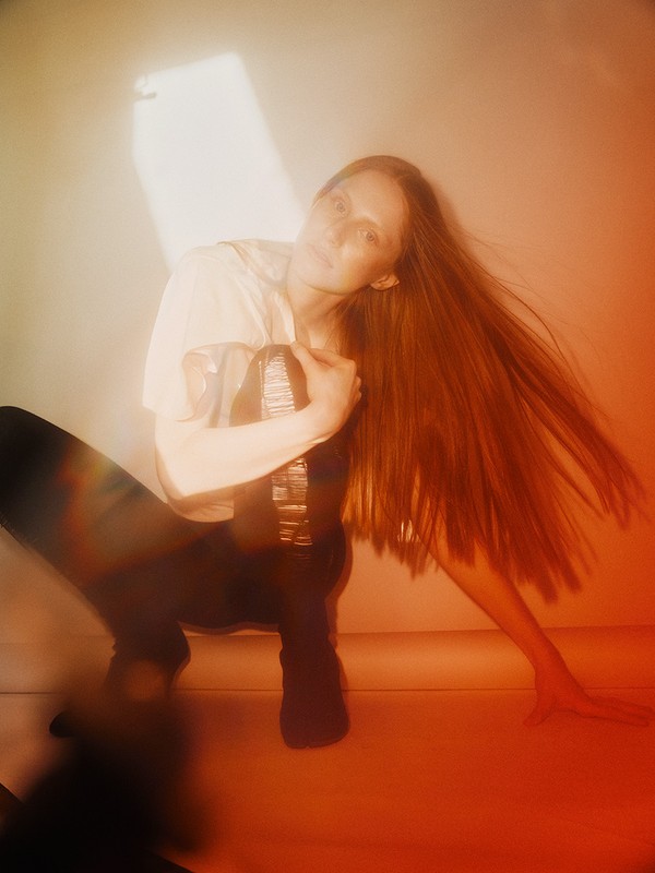 Woman poses in a photo studio, showcasing long reddish-brown hair, wearing black pants and a white top, bathed in soft, diffused lighting.