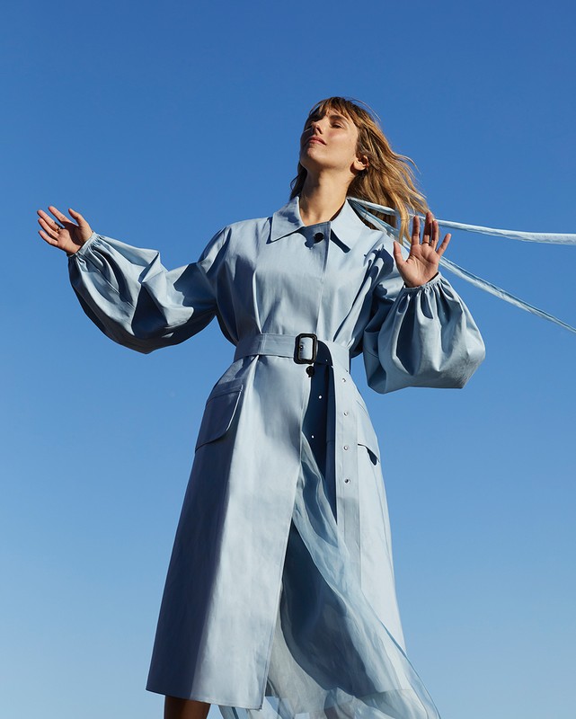 A fashionable adult woman with long hair, wearing a blue trench coat, stands outside with her arms outstretched against a blue sky.