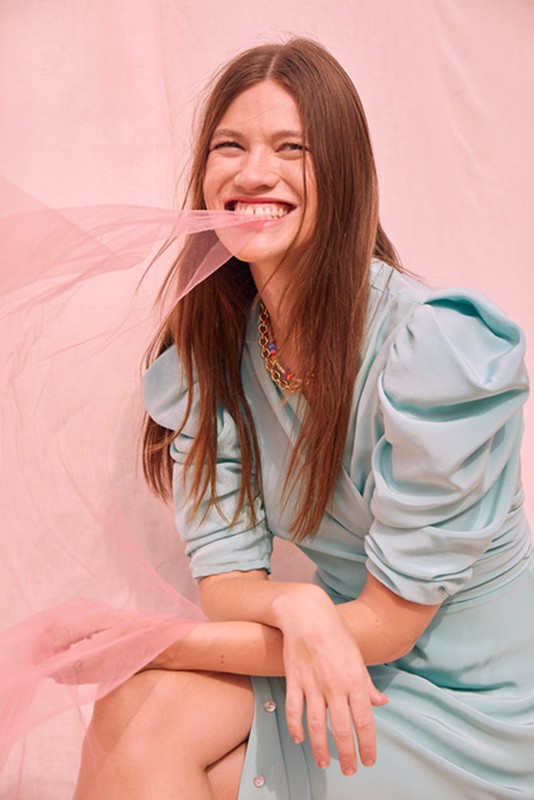 Smiling brunette woman in a blue dress poses in a studio against a pink backdrop with pink fabric in her mouth and hands.