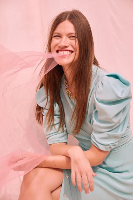 Smiling brunette woman in a blue dress poses in a studio against a pink backdrop with pink fabric in her mouth and hands.