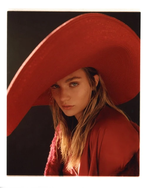 Portrait of a blond female model wearing a large red hat and red outfit against a dark backdrop in a studio setting.