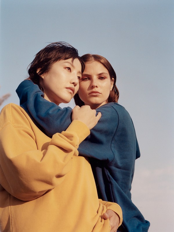 Two women embrace in front of a bright sky, one in yellow sweatshirt, the other in blue, in an outdoor setting. Friendship concept.