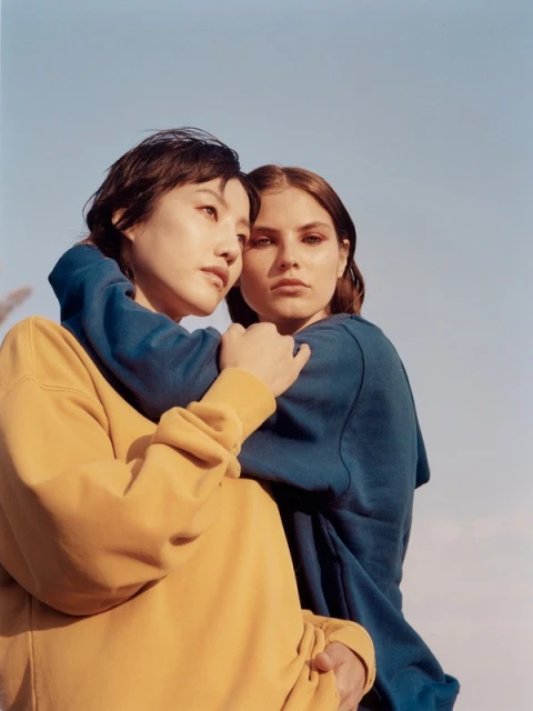 Two women embrace in front of a bright sky, one in yellow sweatshirt, the other in blue, in an outdoor setting. Friendship concept.