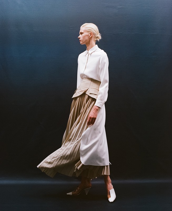 Fashion shot of a woman in a white long-sleeve blouse, khaki corset, and pleated skirt, walking in front of a graduated blue background.