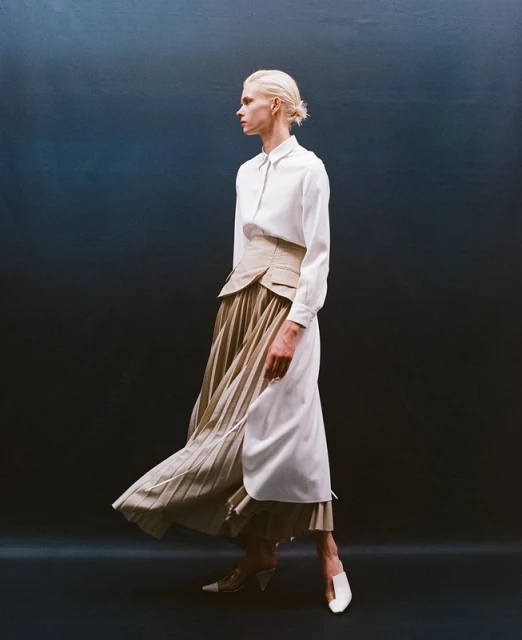 Fashion shot of a woman in a white long-sleeve blouse, khaki corset, and pleated skirt, walking in front of a graduated blue background.