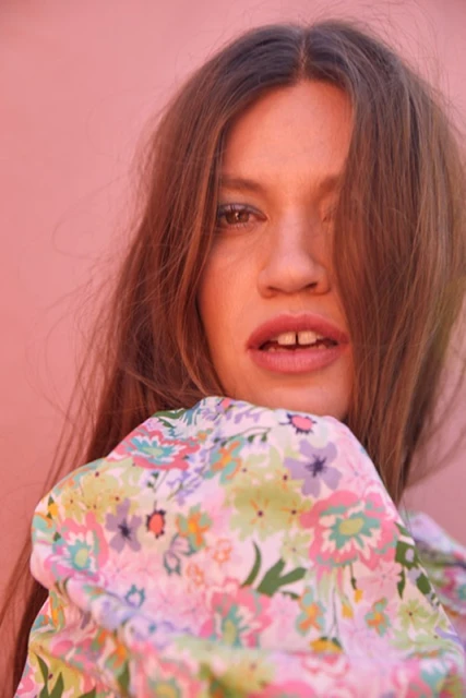Close-up portrait of a young adult female with long brown hair wearing a floral print top against a solid pink background.