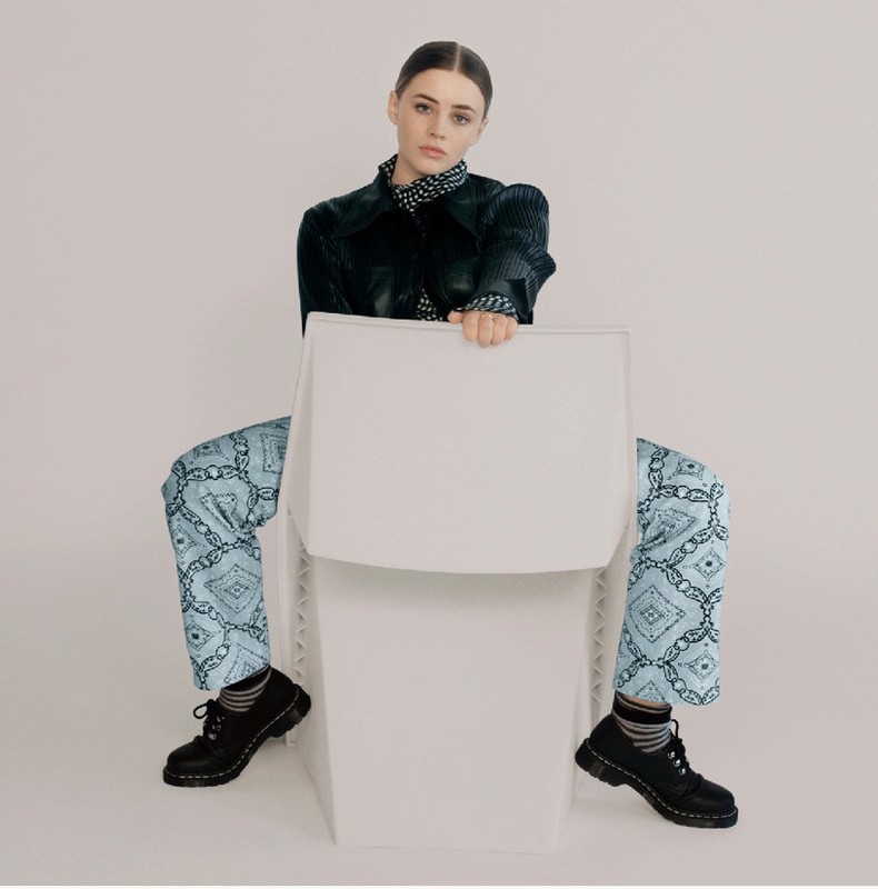 A young woman poses on a white chair in a studio wearing a dark jacket, blue patterned pants, and black lace-up shoes against a white backdrop.