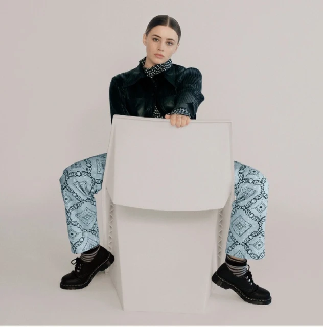 A young woman poses on a white chair in a studio wearing a dark jacket, blue patterned pants, and black lace-up shoes against a white backdrop.