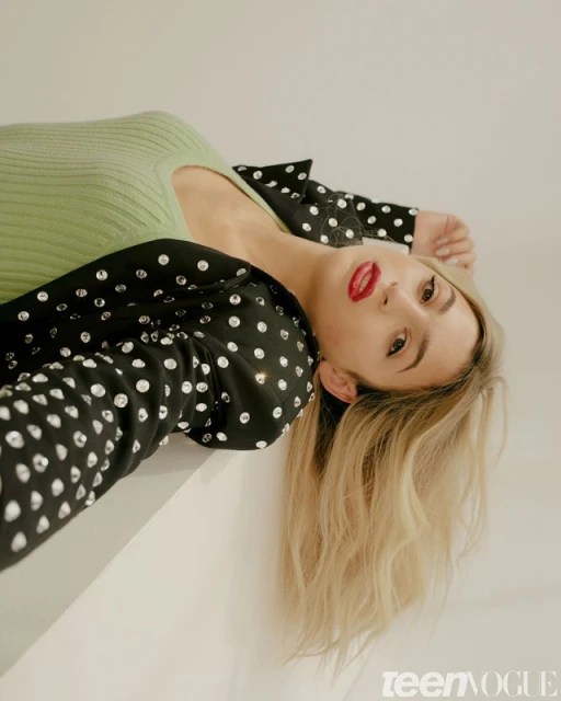 Portrait of a blonde woman in a polka-dot jacket and green top looking at the camera on a neutral background in a studio.