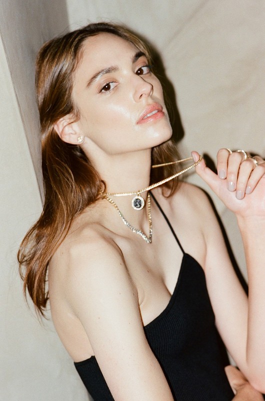 Close-up shot of an adult woman with brown hair posing indoors, wearing a black top and gold necklaces against a neutral background.
