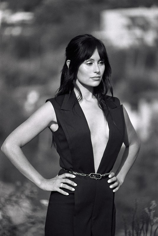Black and white portrait of a woman in a sleeveless black jumpsuit with a chain belt, posing with her hands on her hips outdoors.