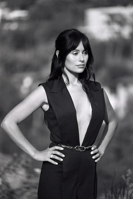 Black and white portrait of a woman in a sleeveless black jumpsuit with a chain belt, posing with her hands on her hips outdoors.