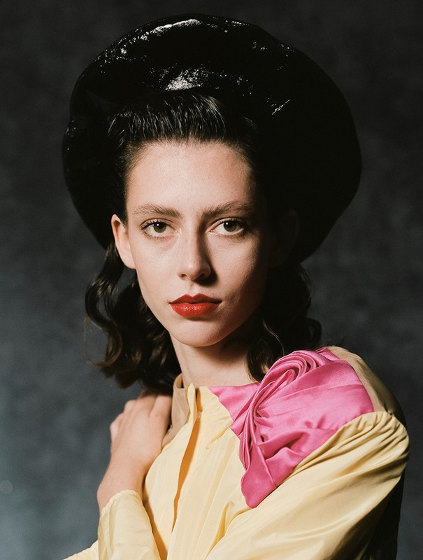 Woman with red lipstick and a yellow top featuring a pink ruffled shoulder detail, wearing a large shiny black hat against a grey background.