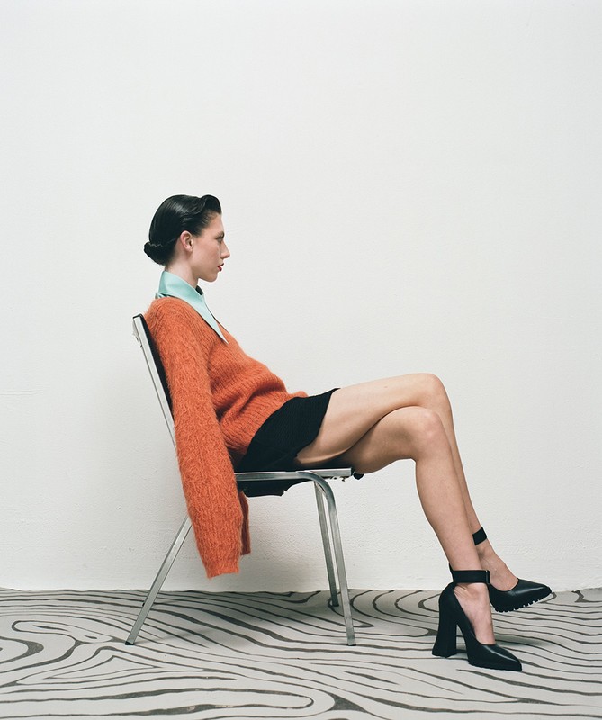 Profile of a woman with dark hair in a bun sitting sideways on a metal chair, wearing an orange sweater and black heels.