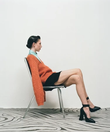 Profile of a woman with dark hair in a bun sitting sideways on a metal chair, wearing an orange sweater and black heels.