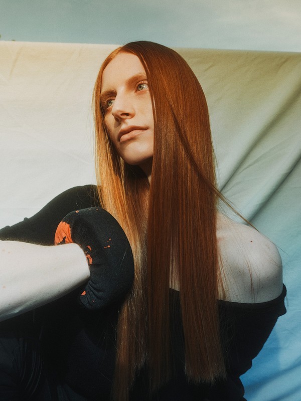 Portrait of a redhead woman with fair skin in a black top posing in front of a light blue and white backdrop with studio lighting.