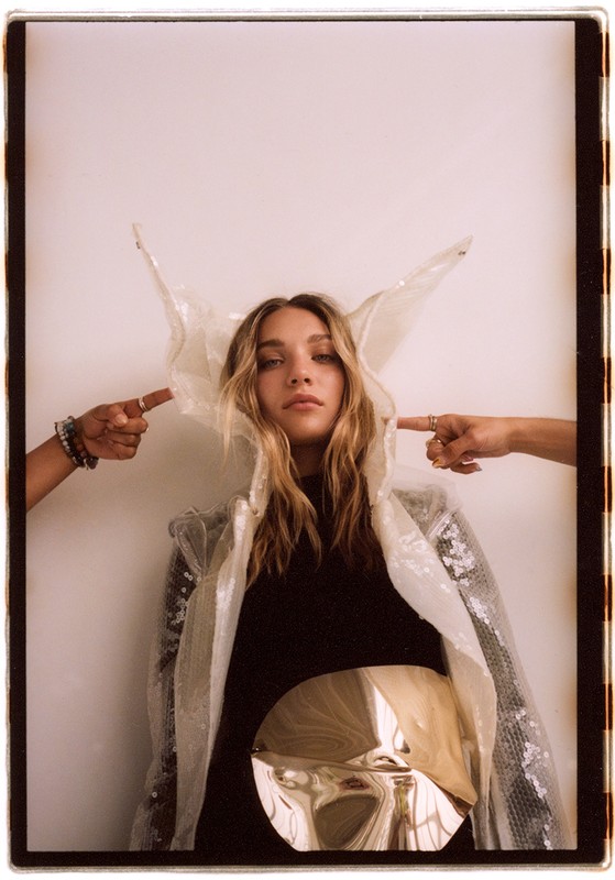 Woman in avant-garde fashion poses in front of a white wall, with two hands pointing at the sculptural headpiece with fingers.