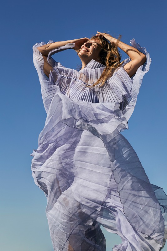 Woman in a lavender pleated dress stands outside with hands on her head, looking up with a blue sky background. Fashion shot.